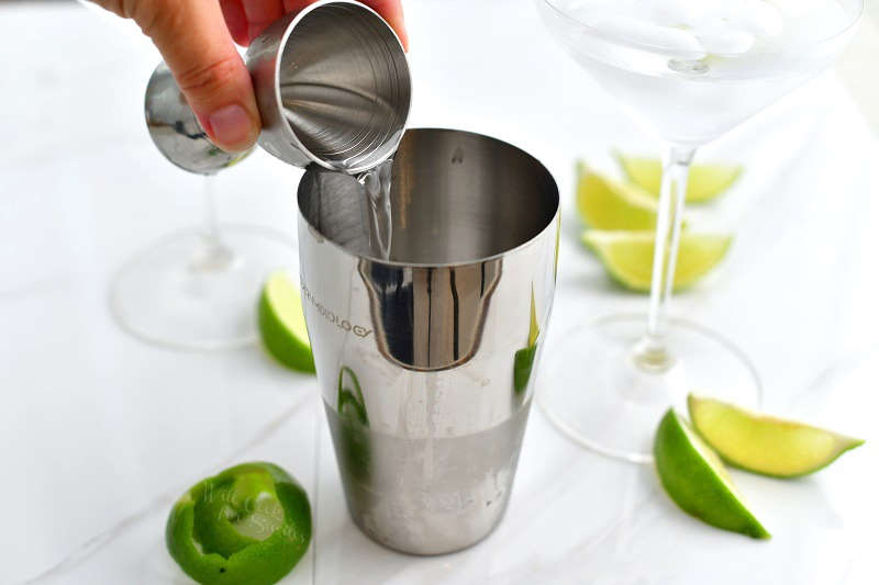 pouring clear vodka into the cocktail shaker out of a silver jigger and martini glass with ice water next to it