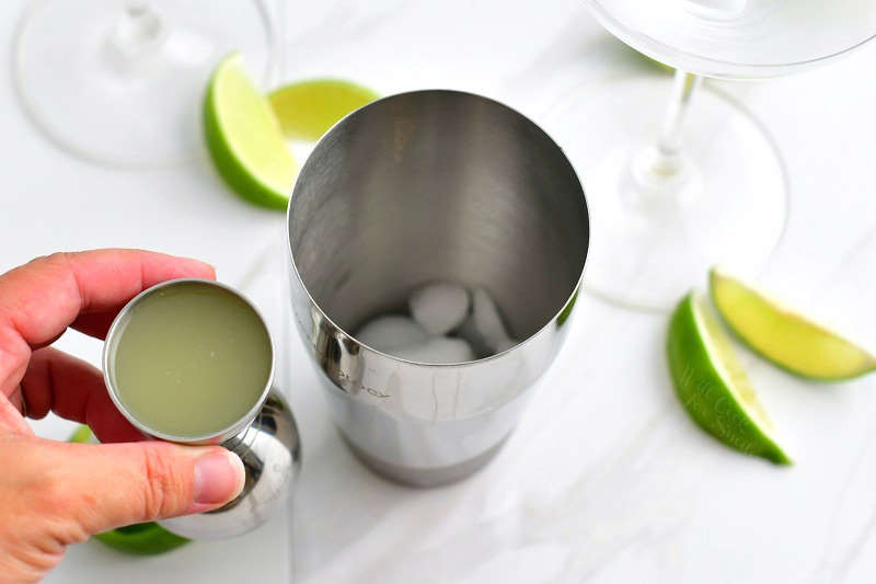 holding fresh lime juice in a metal jigger next to a cocktail shaker with ice and parts of two martini glasses visible