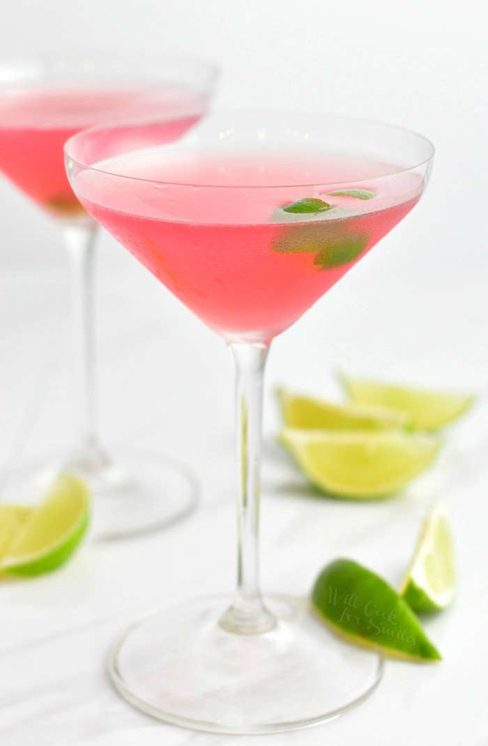side view of the martini glass with pink cocktail and floating lime zest inside, another cocktail on the background and lime wedges on the table