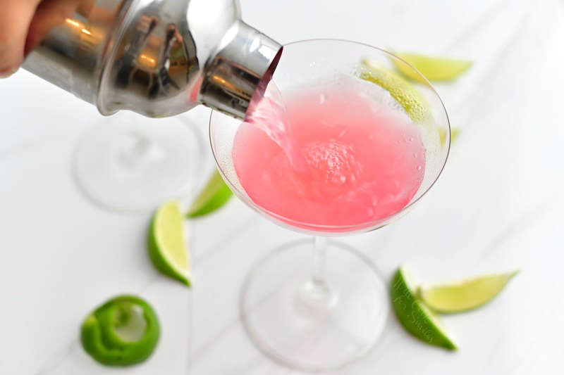 pouring the light pink cosmopolitan cocktail into the chilled martini glass out of a silver cocktail shaker