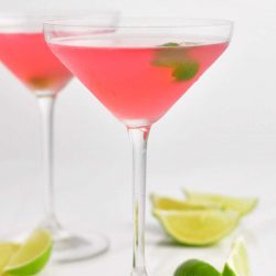 side view of the tall martini glass with with light pink cocktail and a lime zest floating in it and lime wedges scattered around