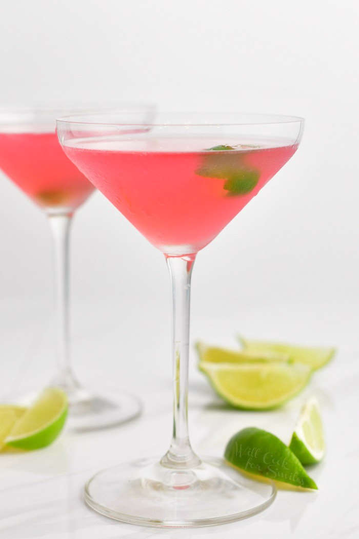 side view of the tall martini glass with with light pink cocktail and a lime zest floating in it and lime wedges scattered around