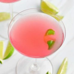 view from the top of the martini glass with light pink cocktail in it and a lime zest floating in the cocktail