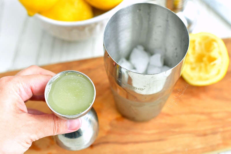 holding silver jigger with lemon juice next to silver cocktail shaker with ice