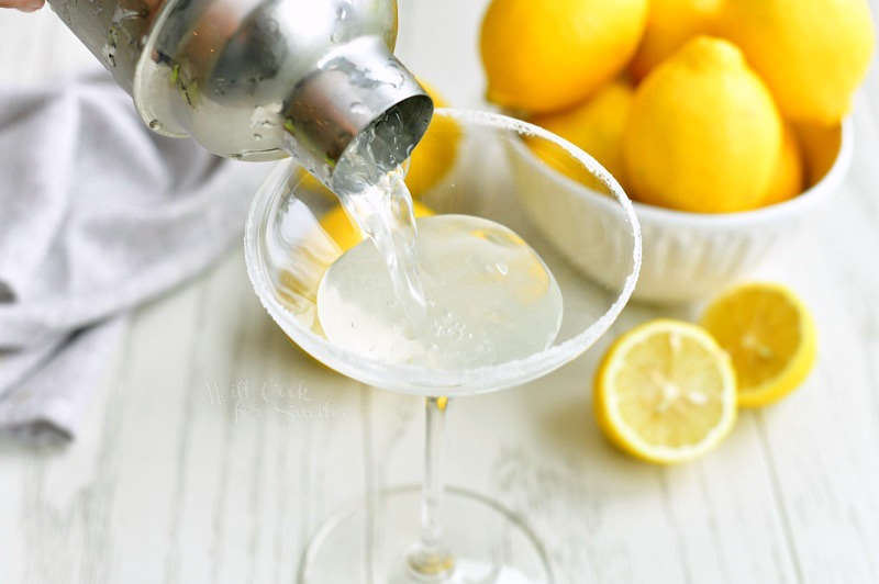 pouring lemon martini into a glass with sugared rim out of a silver shaker with a bowl of lemons next to it