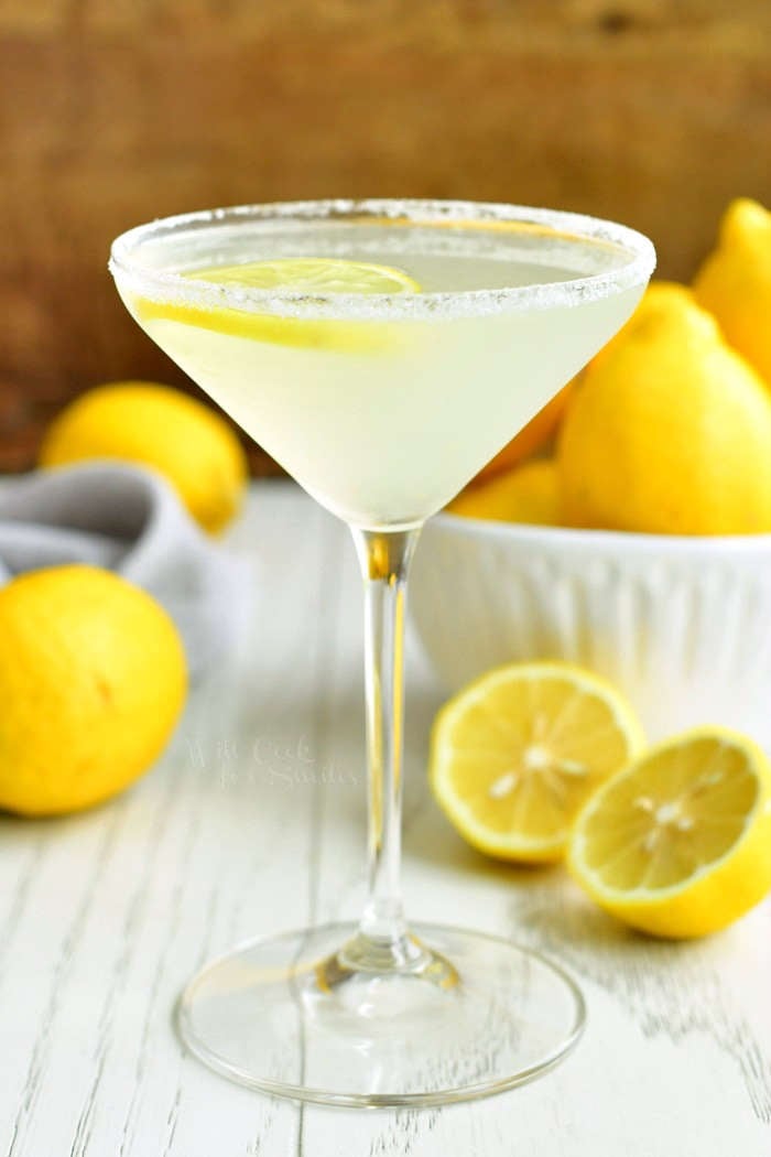 side view of lemon drop martini in a glass with a sugar rim, bowl of lemon next to the glass, and lemon cut in half on the table