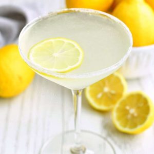 martini glass filled with light yellow cocktail with lemon slice floating in it and sugar on the rim. bowl of lemons next to the glass and some lemon on the table