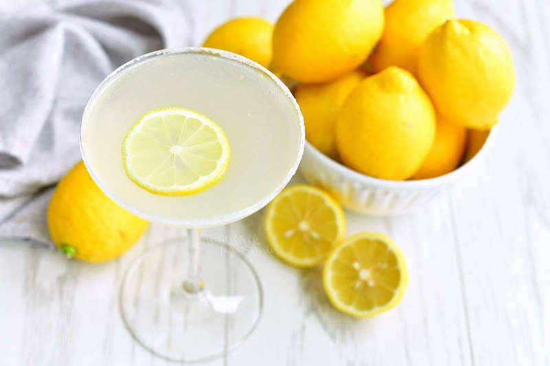 horizontal image of martini glass filled with light yellow cocktail and lemon slice with a bowl of lemons next to it and some lemons on the table