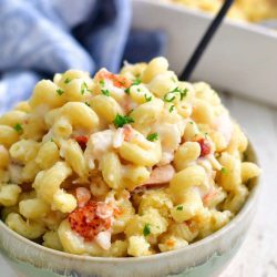 side view of the light blue and tan bowl filled over the top with lobster mac and cheese, a black fork handle visible to the side and baking dish in the background
