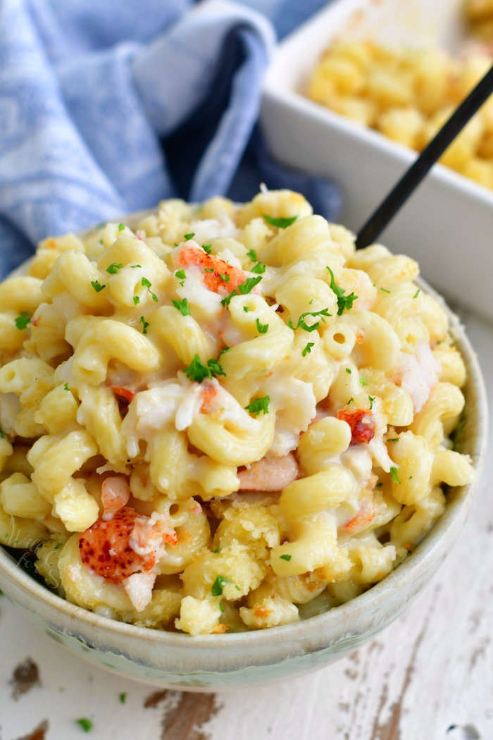 light blue bowl filled over the top with lobster mac and cheese with a little parsley sprinkled on top and black fork handle visible as well as the baking dish in the background