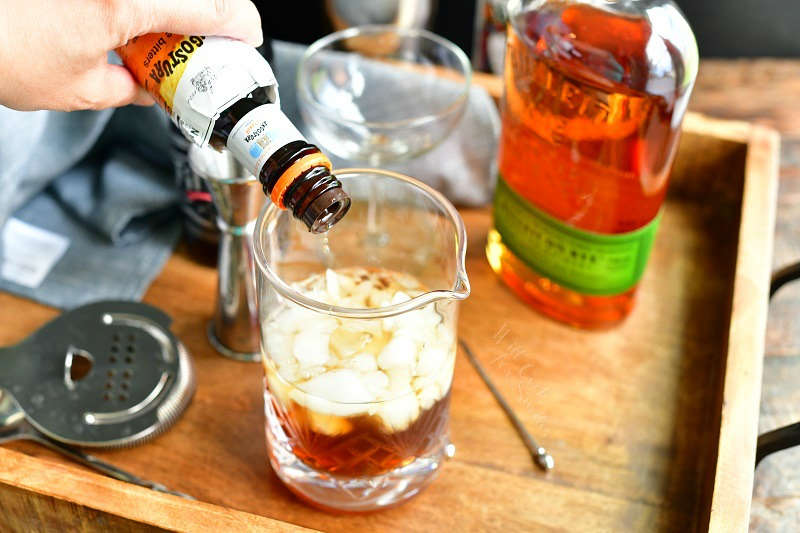 adding dash of bitters into the mixing glass with ice and brown liquors our of a dark brown bottle with bottle of whiskey, glass, jigger, and strainer on the background