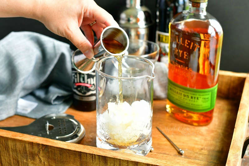 pouring whiskey out of a silver jigger into mixing glass filled with crushed ice. bottle of whiskey on the background with a jar of cherries and sweet vermough