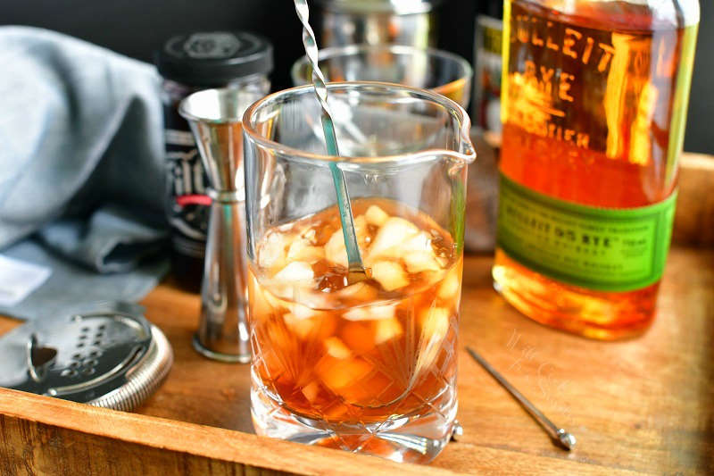 stirring the brown cocktail with ice in a mixing glass using a cocktail long spoon with bottles in the background, strainer and jigger