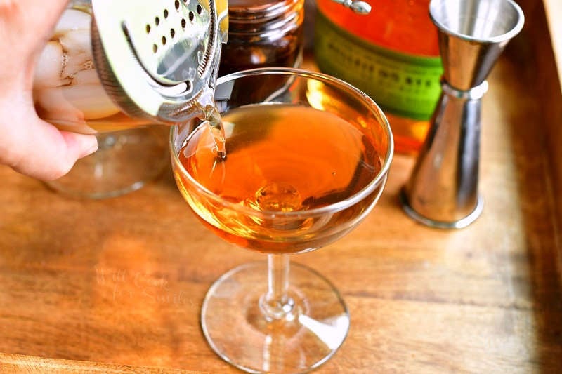 pouring the light brown cocktail into a glass out of the mixing glass covered with silver strainer and a jigger next to the glass