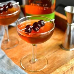 Manhattan cocktail in a glass in the center of the image topped with dark cherries on a silver pock with another glass to the left, a bottle of whiskey on the background and a silver jigger to the right