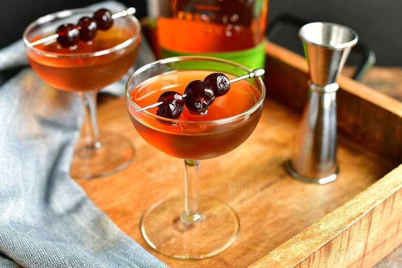 Manhattan cocktail in a glass in the center of the image topped with dark cherries on a silver pock with another glass to the left, a bottle of whiskey on the background and a silver jigger to the right