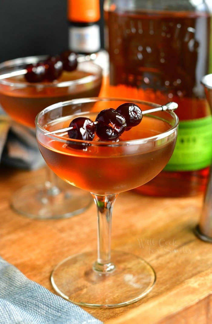 side view of light brown Manhattan cocktail in a glass topped with cherries on a silver pick with another glass on the background and a bottle of rye whiskey