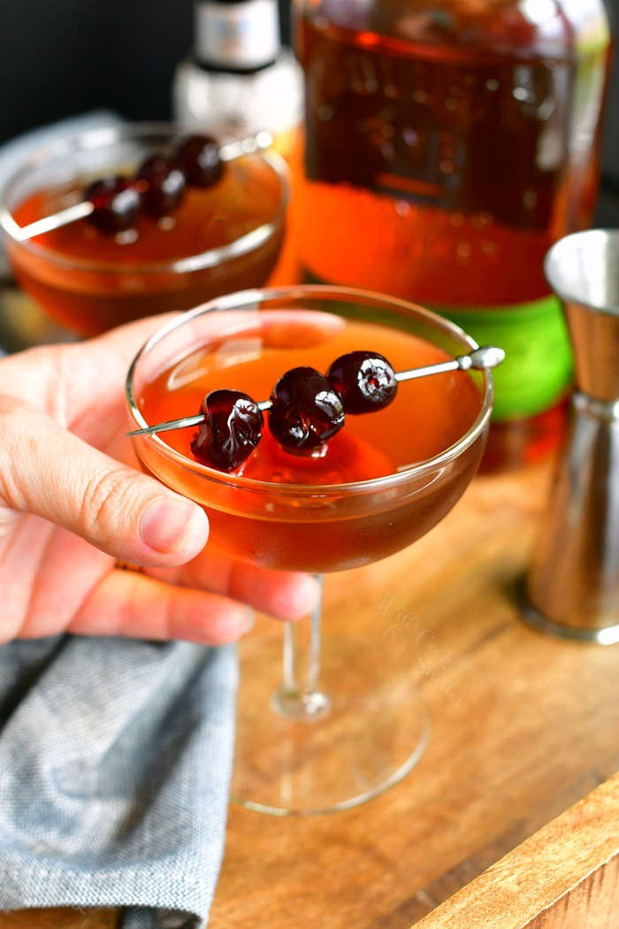 holding a Manhattan cocktail topped with three cherries on a silver pick with another glass on the background and a bottle of whiskey