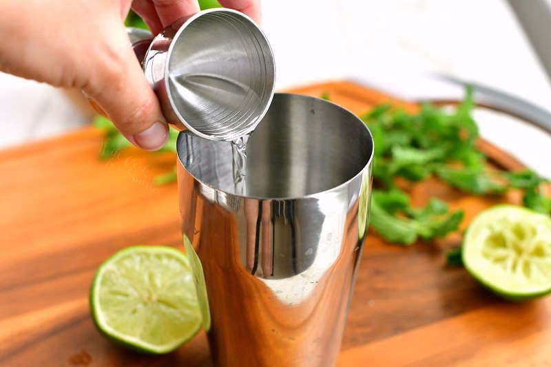 pouring in clear rum from a silver jigger into the silver cocktail shaker with lime halves and mint around