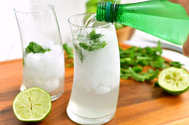 pouring seltzer water from a green bottle into the curved glass with mint rum mixture