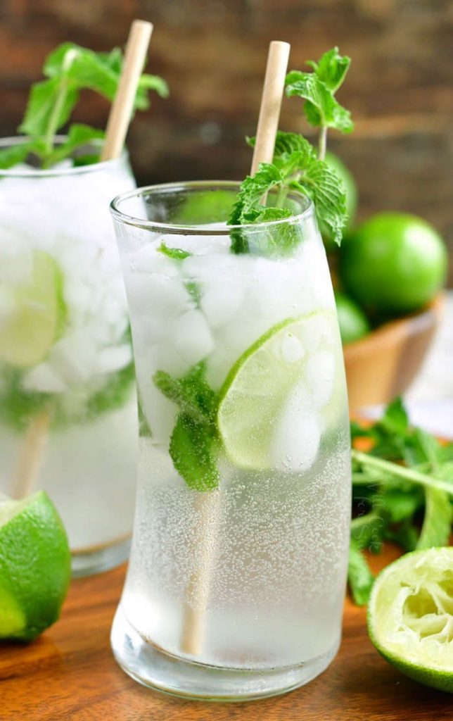 side view of the mijoto cocktail in a curved glass with lime slice inside and mint sprig and paper straw in it, some limes around the glass and another cocktail on the background