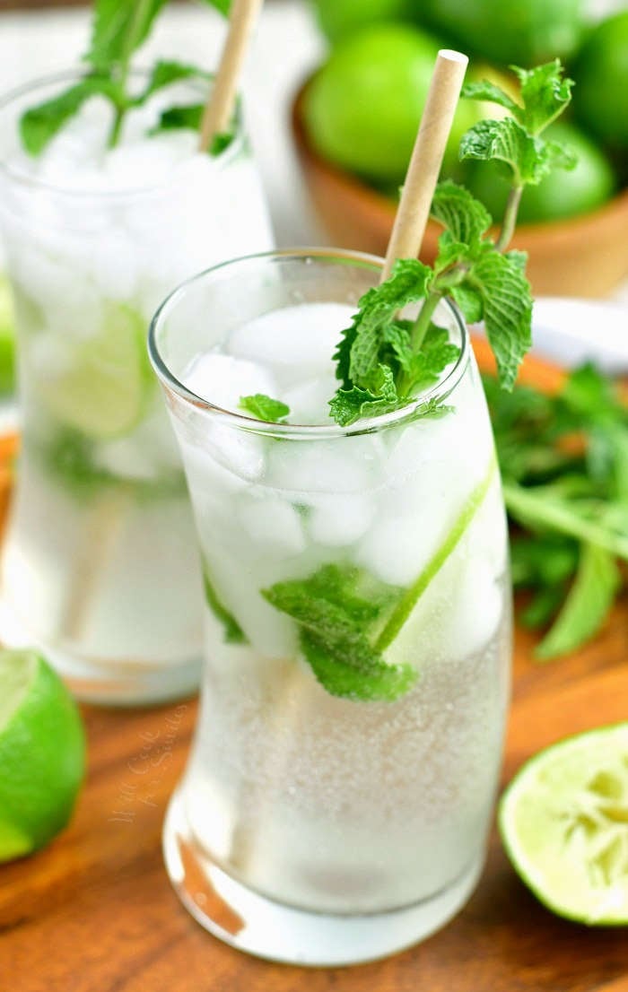 closeup of a mojito cocktail in curved glass with mint sprig and a paper straw in it with another cocktail in the background, limes, and mint on the board