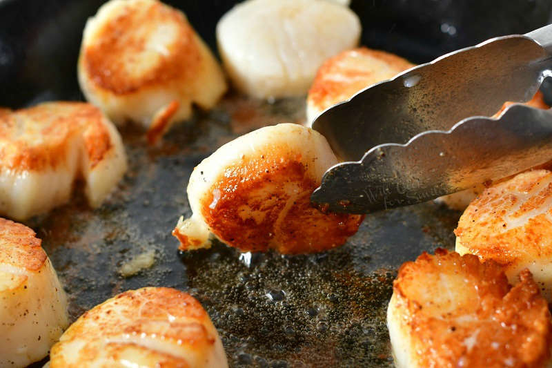 flipping one scallop in a skillet using a metal tongue