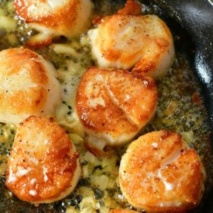 closeup of several seared scallops in a skillet with butter with garlic pieces around