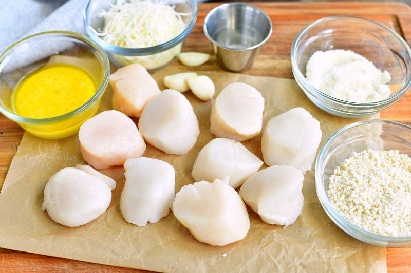 raw scallops on a parchment paper on a wooden board with melted butter, parmesan cheese, salt, garlic cloves, dry parmesan cheese, and Panko crumbs next to them