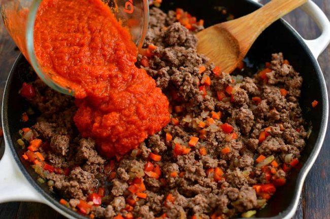 pouring in red tomato sauce into the cooked beef and vegetable mixture in a skillet