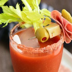 Top of glass shown filled with bloody mary and topped with a speared olive, pickle, cheese and meat garnish as a celery stalk rests on the rim of the glass. A wooden table below with a black and white cloth in the background.