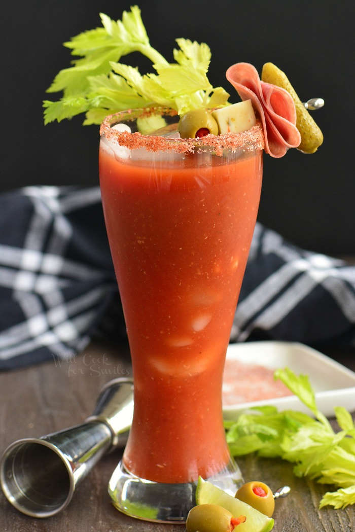 Tall and curved glass filled with red bloody mary and topped with a speared olive, pickle, cheese and meat garnish as a celery stalk rests on the rim of the glass. A wooden table below with a black and white cloth in the background as a cocktail jigger lays on it's side next to the glass at the bottom and additional garnish spear and celery lay next to the glass at the right.