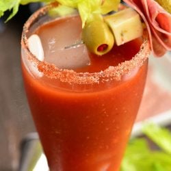 close up of a V8 bloody mary cocktail in tall glass with salted rim and skewer of garnishes including an olive, cheese cube, folded salami, and a pickle