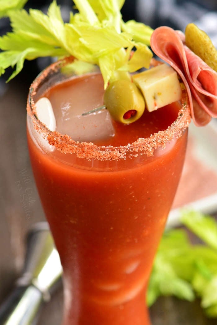 close up of a V8 bloody mary cocktail in tall glass with salted rim and skewer of garnishes including an olive, cheese cube, folded salami, and a pickle