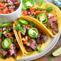 three steak tacos topped with jalapeno rings and pico on a light brown plate with a bowl of pico next to them and some lime slices around