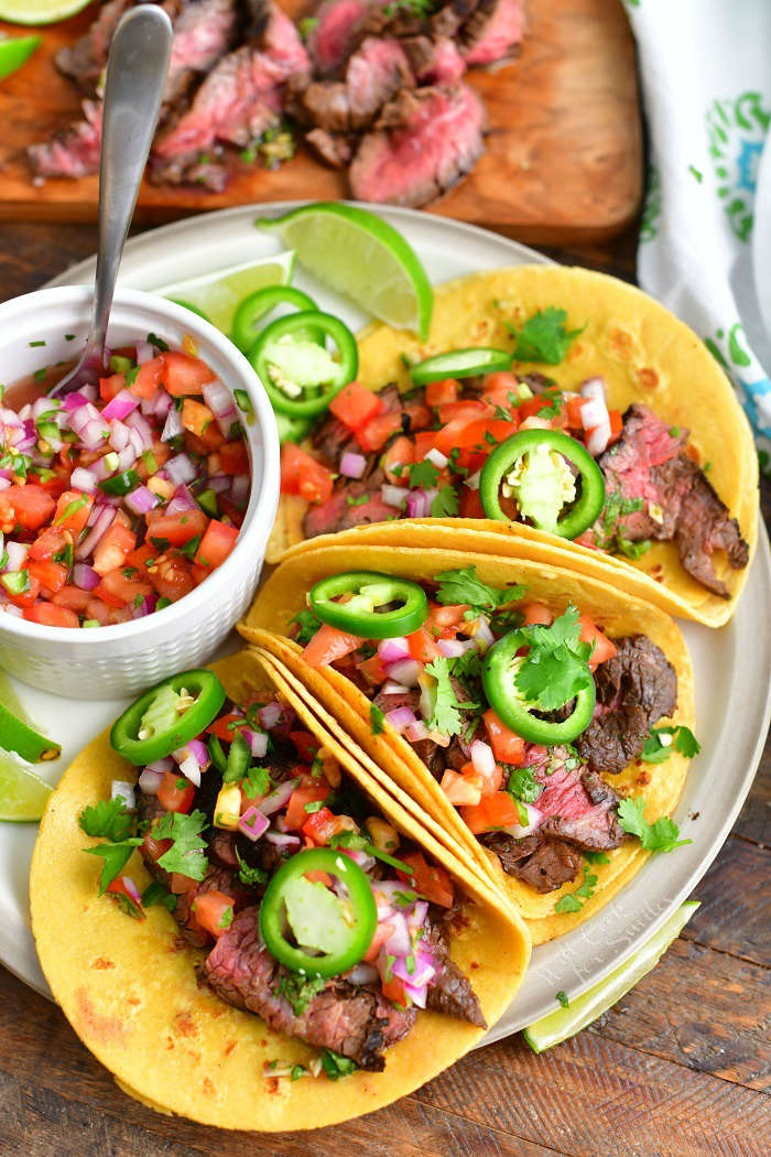 three steak tacos on a plate side by side with a bowl of pico in a plate and sliced steak on a cutting board next to the plate