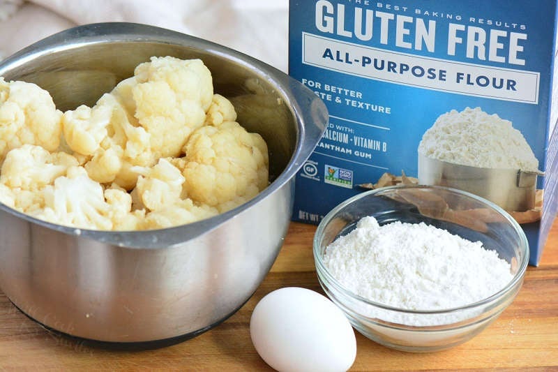 ingredients standing on the cutting board: cooked cauliflower in a metal bowl, small glass bowl of gluten free flour, box of gluten free flour, and 1 egg