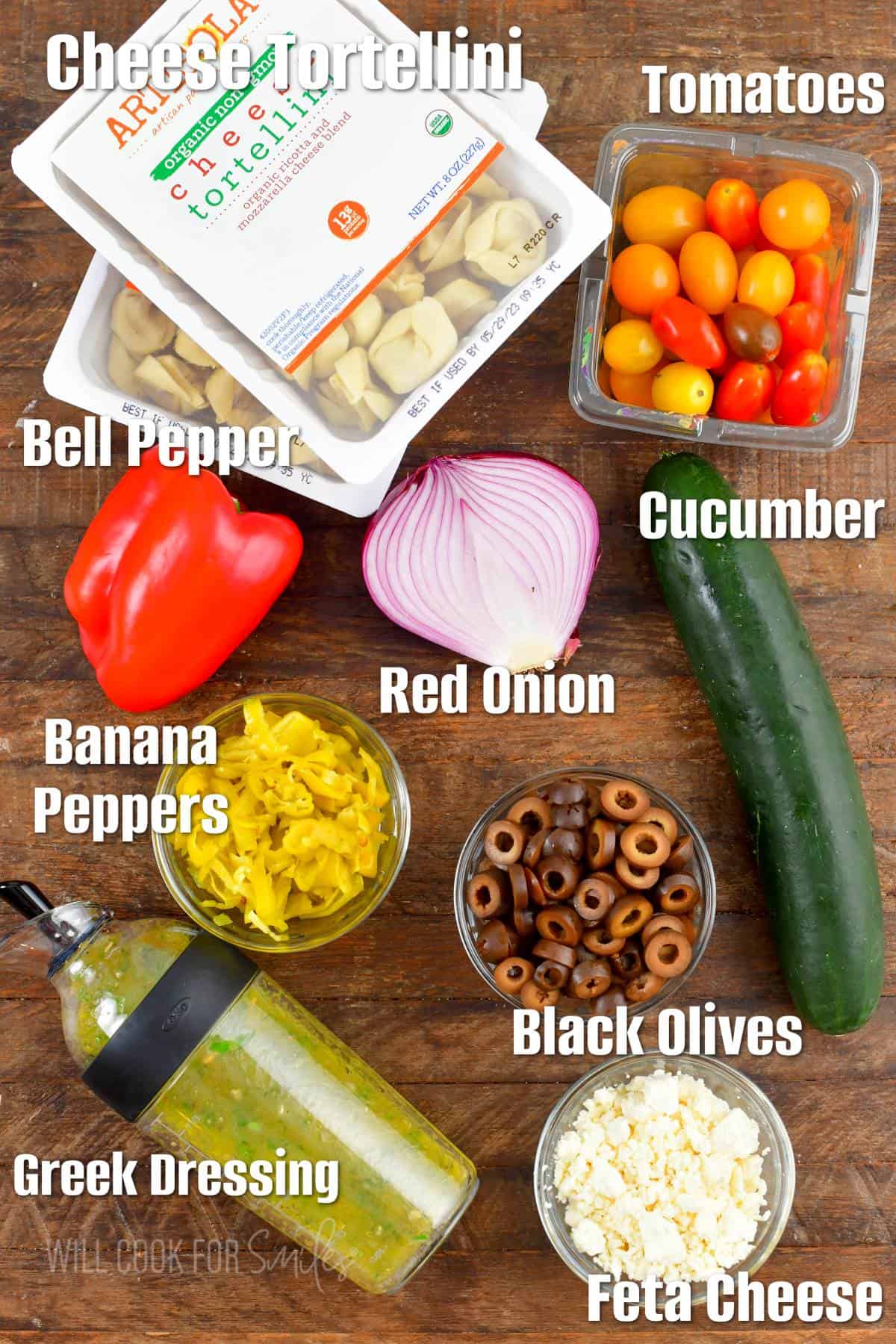 labeled ingredients for Greek tortellini salad on wooden board.