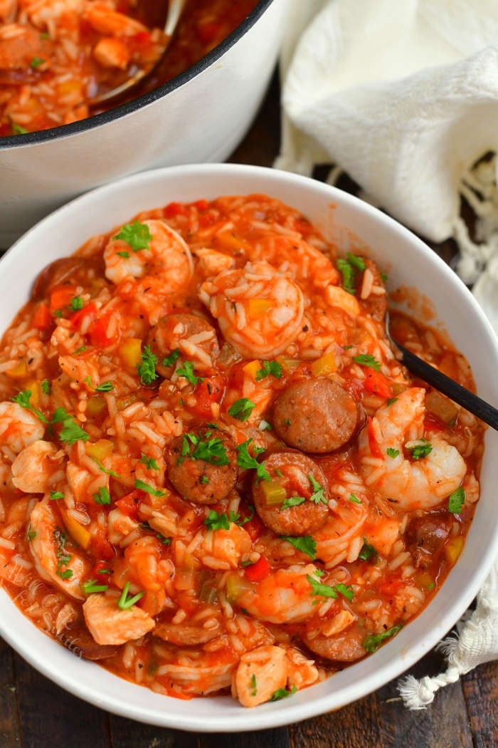 jambalaya in a white bowl with shrimp, sausage, and chicken visible. Black spoon in a bowl and parts of towel around the bowl