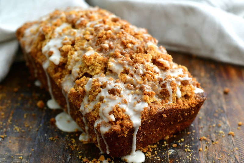 side view of zucchini loaf with crumble topping and white icing drizzled all over the top and dripping off the sides and a grey napkin on the background