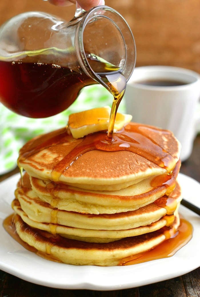 pouring maple syrup over a stack of pancakes out of a glass jar and a cup of coffee behind the plate