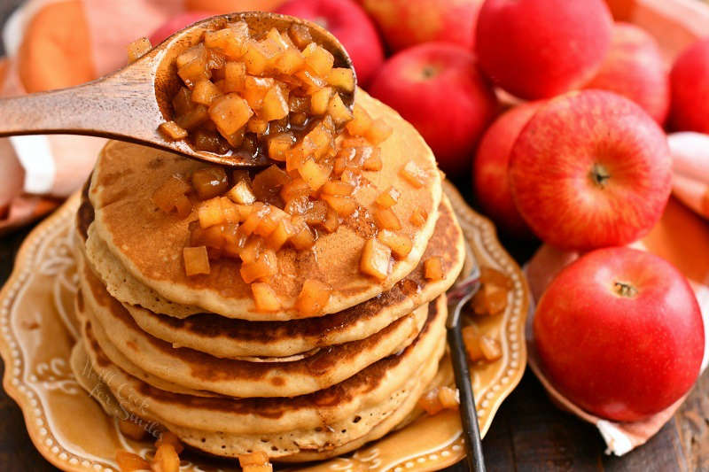 spooning apple pie topping with a wooden spoon over a stack of pancakes