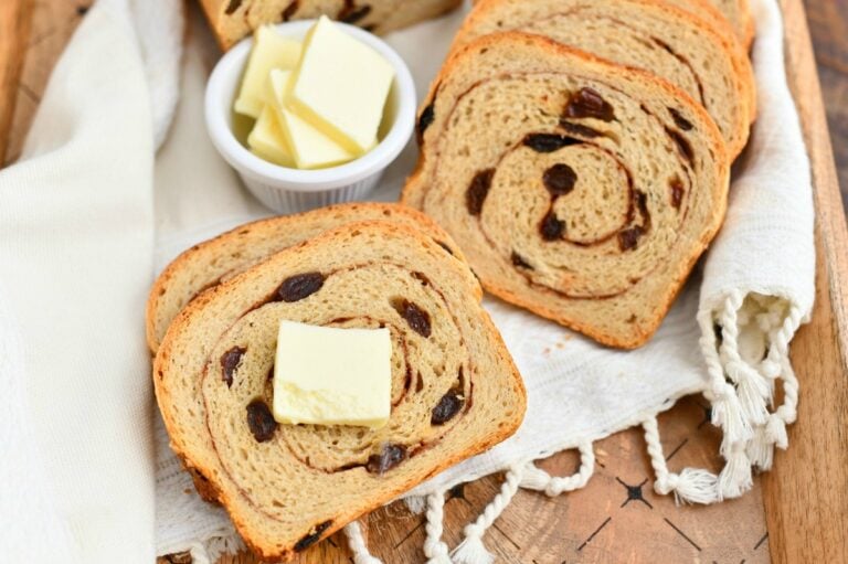 Slices of cinnamon raisin bread are arranged on a wooden board with a small dish of butter. A cloth napkin is partially visible under the bread. Two slices have a pat of butter on top.