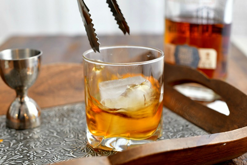 side view: metal tongs dropping ice cube into whiskey cocktail