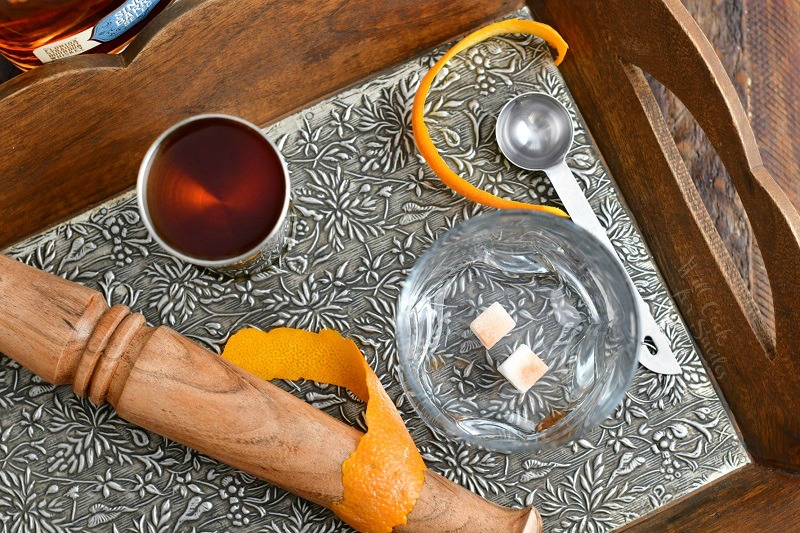 overhead photo of cocktail muddler, jigger of bourbon, sugar cubes, orange peel
