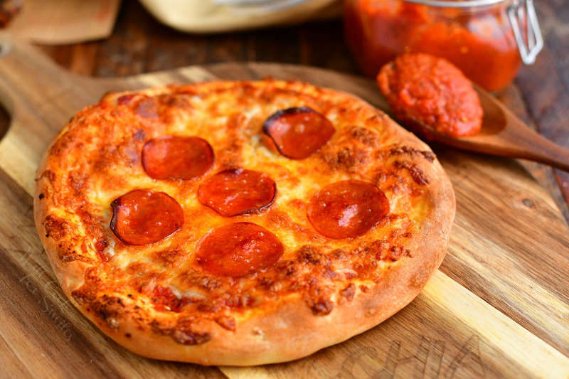 pizza sauce with tomato paste on wooden spoon next to pepperoni pizza