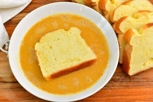 slice of Brioche bread in the egg mixture with more sliced next to the bowl