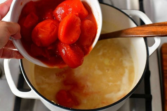 adding canned whole tomatoes to large white pot with chicken stock
