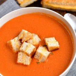 overhead photo: bowl of tomato soup with grilled cheese croutons - grilled cheese sandwich in background