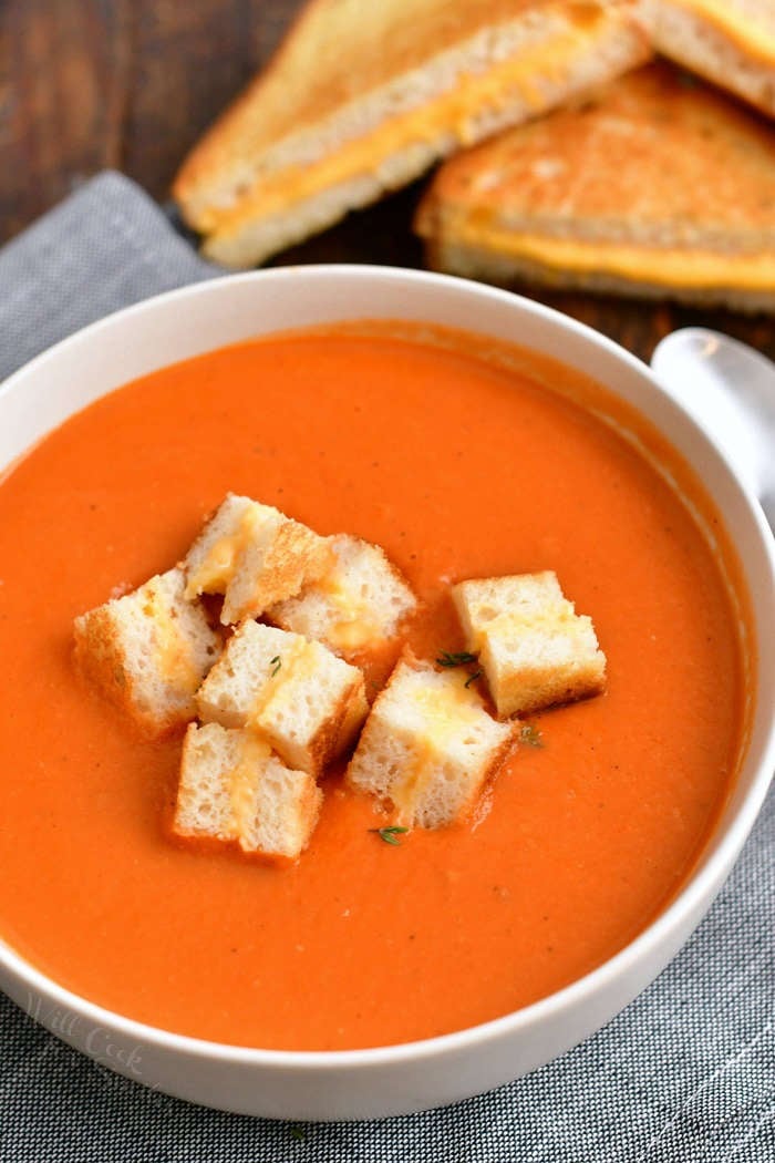 overhead photo: bowl of tomato soup with grilled cheese croutons - grilled cheese sandwich in background